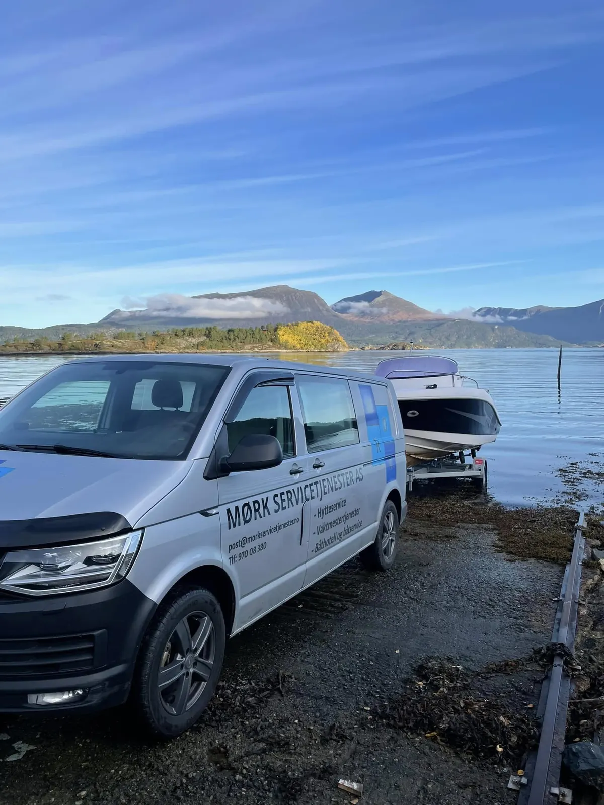 Firmabilen til Mørk Servicetjenester med båt på henger ved en slipp, med fjord og fjell bak