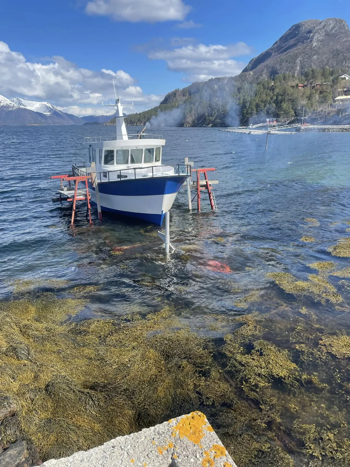 Arbeidsbåt satt opp på bukker i vannkanten med fjord og snødekte fjell bak
